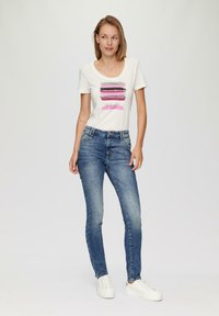 Biała koszulka z krótkim rękawem z kolorowymi paskami i napisem "BĄDŹ ŚWIADOMY", w połączeniu z niebieskimi obcisłymi dżinsami i białymi trampkami.