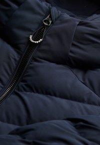 Navy pufferjack met gequilte textuur, voorzien van een zwarte rits en trekband. De materialen zijn strak en glad, wat zorgt voor isolatie.