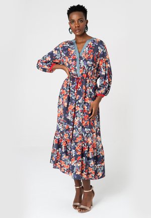 Kobieta ubrana w długą sukienkę w kwiaty w kolorach pomarańczowym, białym i czerwonym na ciemnym tle, stojąca z jedną ręką na biodrze i beżowymi szpilkami.