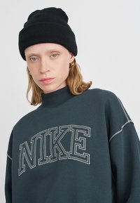 Tumši teālī zēna ar baltu "NIKE" logo, augsts apkakles un gaiša šuvuma. Valkāta ar melnu adītu cepuri. Gluda tekstūra, ikdienišķs dizains.