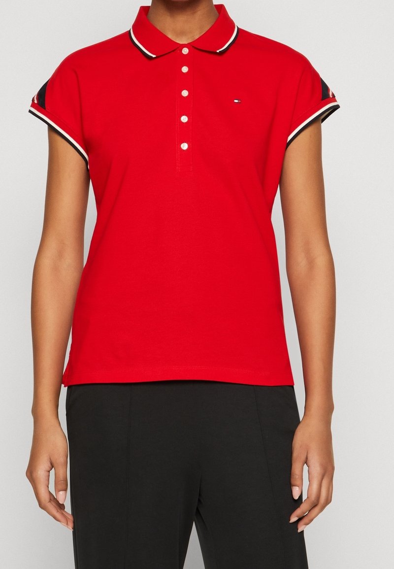 Femme portant un polo rouge à manches courtes avec des boutons blancs et un pantalon noir, debout devant un fond clair uni.