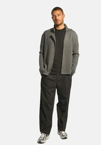 Grå kofta över svart t-shirt, kombinerad med svarta byxor. Modellen har på sig vita och svarta sneakers och står mot en vit bakgrund.