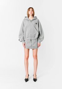 Sudadera gris con cremallera y capucha, de corte oversized, con detalle de logo, combinada con una minifalda gris a juego, complementada con tacones de aguja negros.