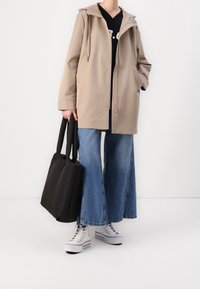 Giacca beige con cappuccio e zip, abbinata a jeans larghi in denim chiaro e sneakers bianche alte. Una borsa tote nera è tenuta in una mano.