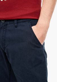 Dospělá osoba má pravou ruku položenou v přední kapsy tmavě modrých kalhot, na sobě má červenou košili.