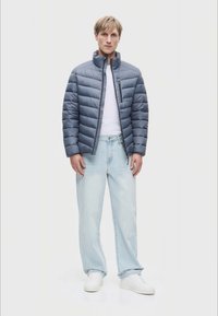 Párnázott sötétkék dzseki átlós varrással, cipzárral és magas gallérral, fehér póló és világoskék farmer felett viselve, fehér sportcipővel párosítva.