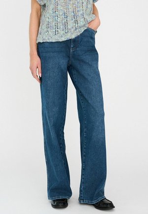 Széles szárú, kék farmernadrág egyenes szabással, finom kifakulással, öt zsebes kialakítással és kontrasztos varrással. Párban egy kötött felsővel viselve.