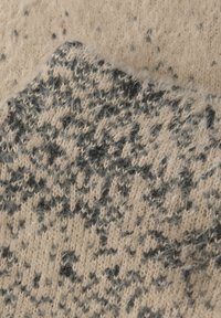 Stickad textil i krämfärgad och grå, med en mjuk, fluffig textur och ett prickigt mönster. Närbild som framhäver vävstrukturen.