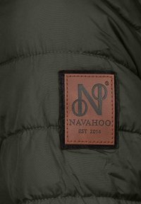 Mörkgrön puffjacka i tyg med kviltad textur. Har en fyrkantig brun läderetikett med svart logotyp och texten "NAVAHOO EST 2014."