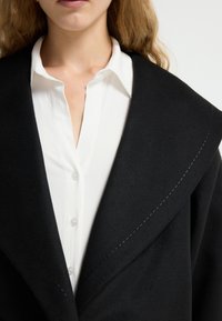Zwarte wollen jas met een brede kraag, voorzien van zichtbare stiksels, gelaagd over een witte gestructureerde blouse met knoopsluiting.