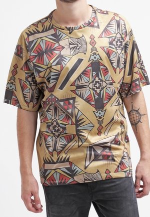 Chemise à manches courtes présentant un motif géométrique en noir, rouge et teal sur un fond beige. Le tissu semble léger avec une coupe ample.