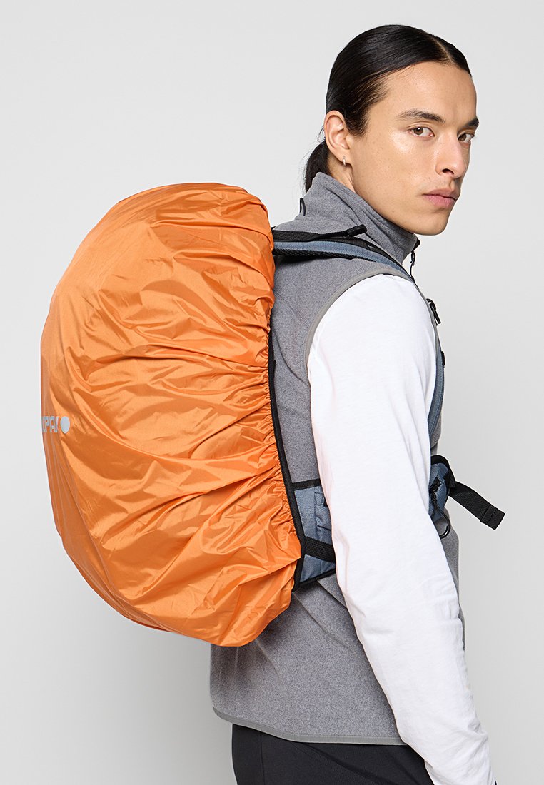 Orangen wasserfester Schutz über einem grauen Rucksack, präsentiert an einem Modell, das eine graue Weste und ein weißes Langarmshirt trägt.