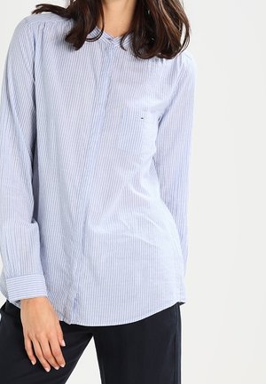 Kvinde iført en lyseblå og hvid, vertikalt stribet langærmet skjorte med brystlomme og sorte bukser, stående mod en ensfarvet baggrund.