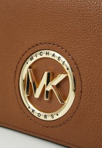 Borsa in pelle marrone con una texture, caratterizzata da un logo circolare dorato con "Michael Kors" e le iniziali intrecciate "MK" al centro.