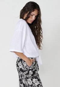 Camicia bianca di lino a maniche corte con vestibilità comoda, abbinata a pantaloni neri con motivo floreale e dettagli con tasche.