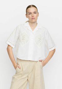 Weißes Cropped Shirt mit gestickten Akzenten und kurzen Ärmeln, mit Kragen und Knopfverschluss, kombiniert mit beigen weit geschnittenen Hosen.