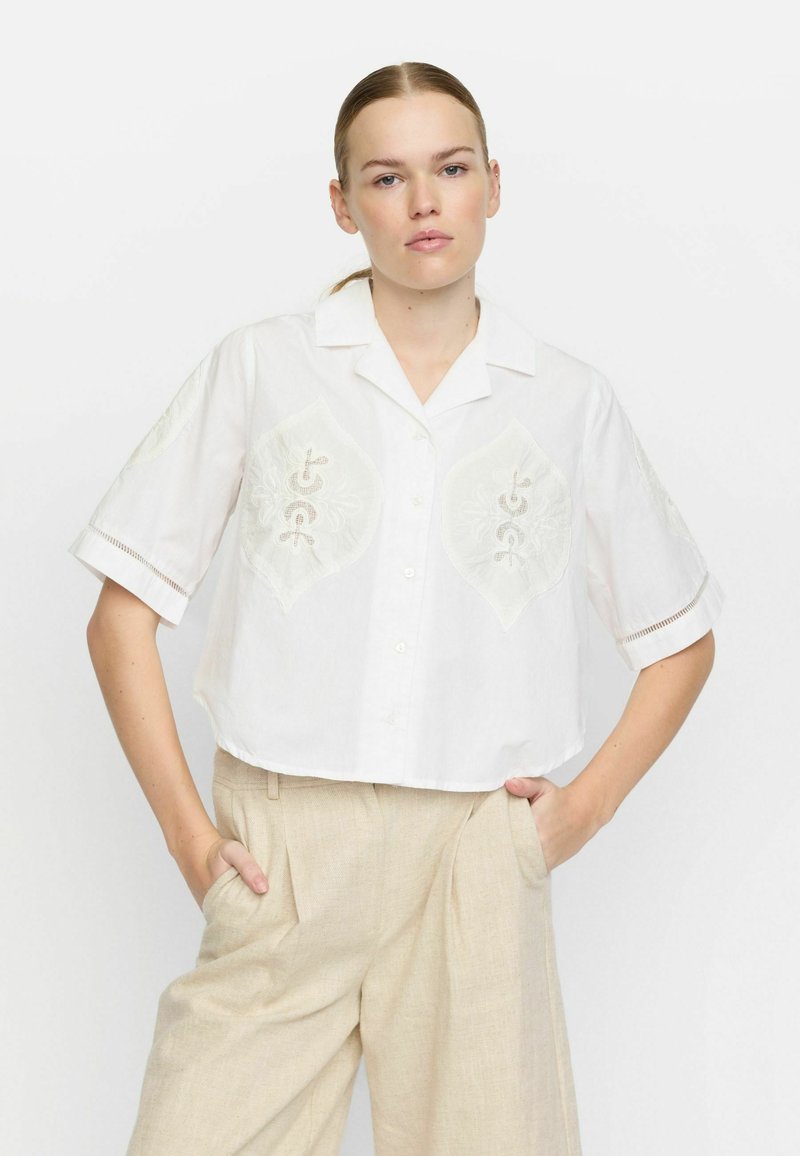 Weißes Cropped Shirt mit gestickten Akzenten und kurzen Ärmeln, mit Kragen und Knopfverschluss, kombiniert mit beigen weit geschnittenen Hosen.