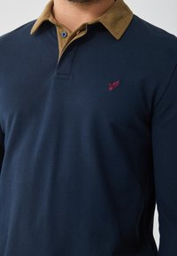 Marineblå langermet polo skjorte med en beige kordfløyelskrage, som har en liten rød fugl brodert på brystet og en to-knapps stolpe.