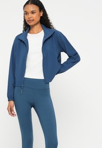 Jacket atlética azul com fecho éclair à frente e punhos elásticos, combinada com leggings de cintura alta em um material liso e elástico.