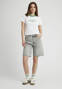 T-shirt blanche coupée avec encolure verte et inscription "AUTHENTIC". Shorts en denim gris à la coupe décontractée. Chaussures blanches avec accents noirs et chaussettes rayées vertes.