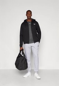 Man met zwarte rits-hoodie, grijs T-shirt, lichtgrijze trainingsbroek, witte sneakers, met een zwarte sporttas in een studioomgeving.