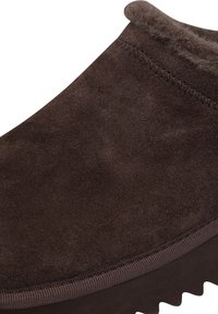 Zapato deslizante de ante marrón con una superficie texturizada, borde superior redondeado y suela de goma con patrones de agarre. Forro suave visible en la apertura.