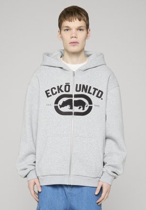 Młoda osoba ubrana w jasnoszarą bluzę z kapturem na suwak z logo „ECKŌ UNLTD.” oraz grafiką nosorożca, w niebieskich dżinsach, stojąca na jednolitym tle.
