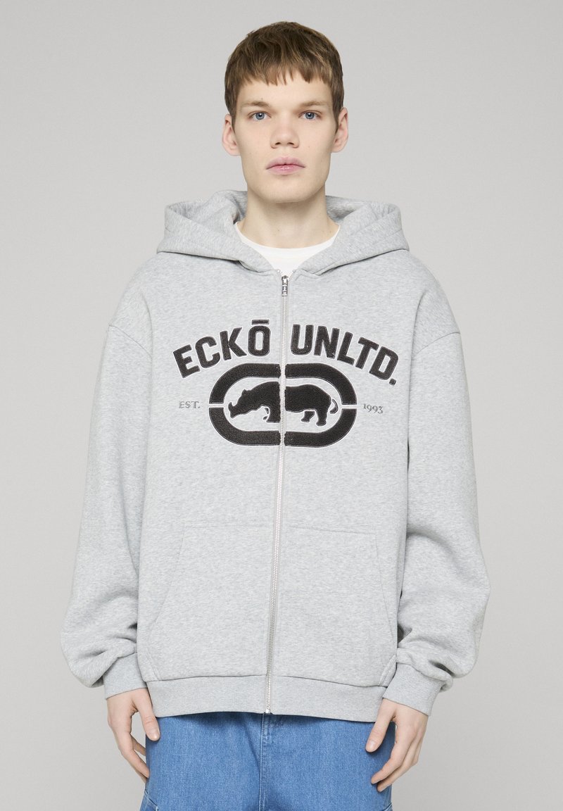 Jeune personne portant un sweat à capuche zippé gris clair avec le logo "ECKŌ UNLTD." et un motif de rhinocéros, associée à un jean bleu, debout devant un fond uni.