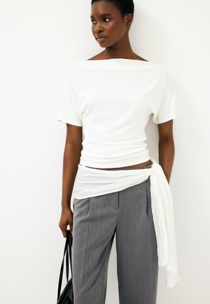 Frau trägt weißes Kurzarm-Top mit transparentem, gebundenem Wickeldetail und graue Hochbundhose, hält eine schwarze Handtasche.