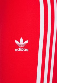 Röda sportleggings tillverkade av elastiskt tyg. Har tre vita ränder längs sidorna och en vit Adidas-logotyp på låret.