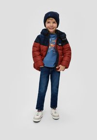 Rote und marineblaue Daunenjacke mit Kapuze, blaues T-Shirt mit Grafik, blaue Jeans und weiße Sneakers mit grauen Akzenten. Der Junge trägt eine marineblaue Mütze.