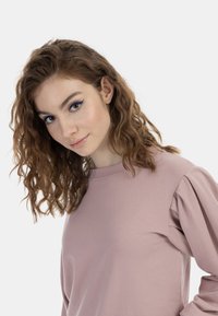 Felpa rosa chiaro con maniche lunghe, caratterizzata da dettagli spalline a sbuffo. Texture liscia, design con collo rotondo e vestibilità casual.