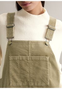 Oliwkowe sztruksowe ogrodniczki z regulowanymi metalowymi ramiączkami, z przednią kieszenią i pionową prążkowaną fakturą, noszone na kremowym swetrze.