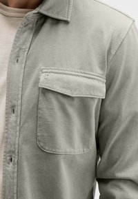 Hellgraues Hemd mit Knöpfen, gefertigt aus weichem Stoff, mit einer einzelnen Brusttasche und einem kragenförmigen Ausschnitt. Mit dezenten Stickdetails strukturiert.