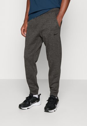 Mężczyzna w ciemnoszarych spodniach dresowych Nike z małym czarnym logo, czarno-białych butach Nike i niebieskiej koszuli, stojący z jedną ręką w kieszeni.