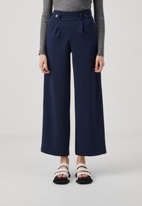 Personne portant un pantalon large bleu marine avec des détails à boutons et un haut gris côtelé à manches longues, associé à des sandales blanches et noires.