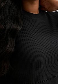 Frau, die ein schwarzes, geripptes Langarmshirt trägt, mit langen, dunklen Haaren, die über ihre Schulter fallen, und einer Hand, die nahe am Hals ruht.