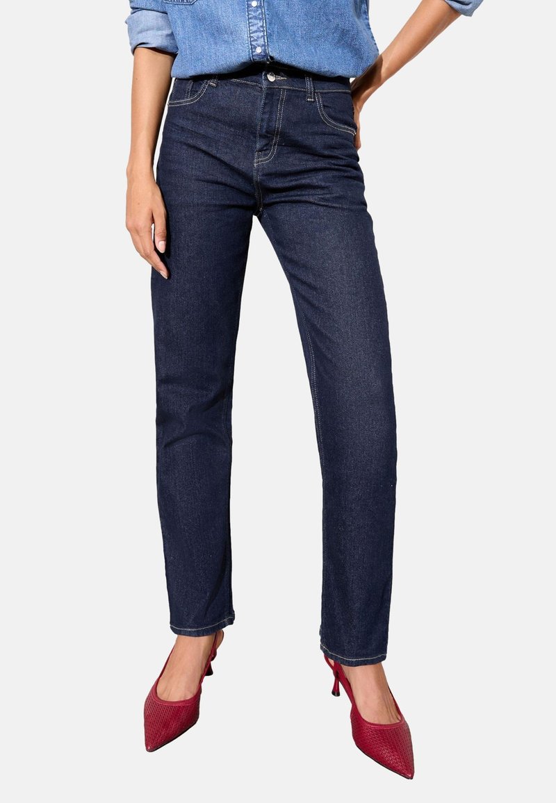 Frau trägt dunkelblaue Jeans, ein hellblaues Jeanshemd und rote spitze High Heels und steht vor einem einfarbigen Hintergrund.
