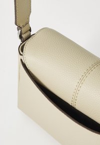 Beige lærveske med rund form, med teksturert overflate, brune kanter og avtakbar stropp. Åpen lomme-design foran.