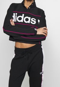 Svart kort sweatshirt med vit "adidas"-logotyp och rosa detaljer, kombinerat med svarta mjukisbyxor med vita sidstroppar och en logotypapplikation.