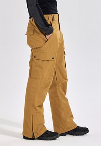 Tan cargobroek met een relaxte pasvorm, voorzien van meerdere zakken, ritsdetails en een duurzame stof. G gedragen met zwarte schoenen.