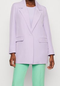 Blazer lilas avec un col cranté, une fermeture à un bouton et deux poches avant, assorti à un pantalon vert menthe. Tissu à texture lisse.