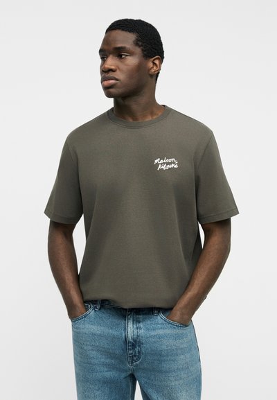 Νέος άνδρας με πράσινη ελιά κοντομάνικη μπλούζα Maison Kitsuné με λευκό λογότυπο στο στήθος και μπλε τζιν, με τα χέρια στις τσέπες, κοιτάζοντας αριστερά.