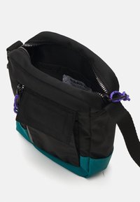 Svart tyg axelväska med en petrolblå botten. Har en dragkedjestängning, ett externt fack och lila dekorativa detaljer. Intern etikett synlig.