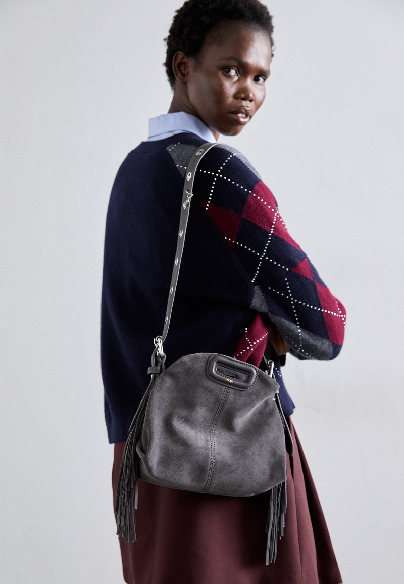 Frau trägt einen dunkelblauen und burgunderfarbenen Argyle-Pullover mit einer grauen Wildleder-Fransen-Handtasche über der Schulter und blickt über ihre Schulter zurück.