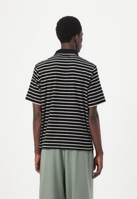 Black and white striped short-sleeve polo shirt with a collar, displayed from the back, paired with loose green trousers.