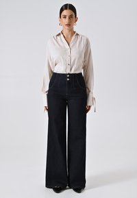 Lichtbeige button-up shirt met een kraag, gecombineerd met hooglopende donkere denim broek met wijde pijpen, voorzien van zijzakken en gouden knopen.