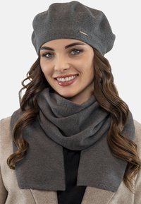 Szürke kötött beret, sima textúrával és kis fém logóval, párosítva egy hozzáillő szürke sállal, amely puha, redőzött kialakítású.