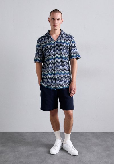 Uomo in piedi contro un muro grigio che indossa una camicia a maniche corte con motivo a zigzag, pantaloni corti blu navy, calze bianche e sneakers bianche.