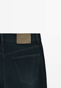 Jeans en denim bleu foncé avec une texture lisse, dotés d'une étiquette en cuir rectangulaire portant la mention "Massimo Dutti" sur la ceinture et d'une poche arrière.